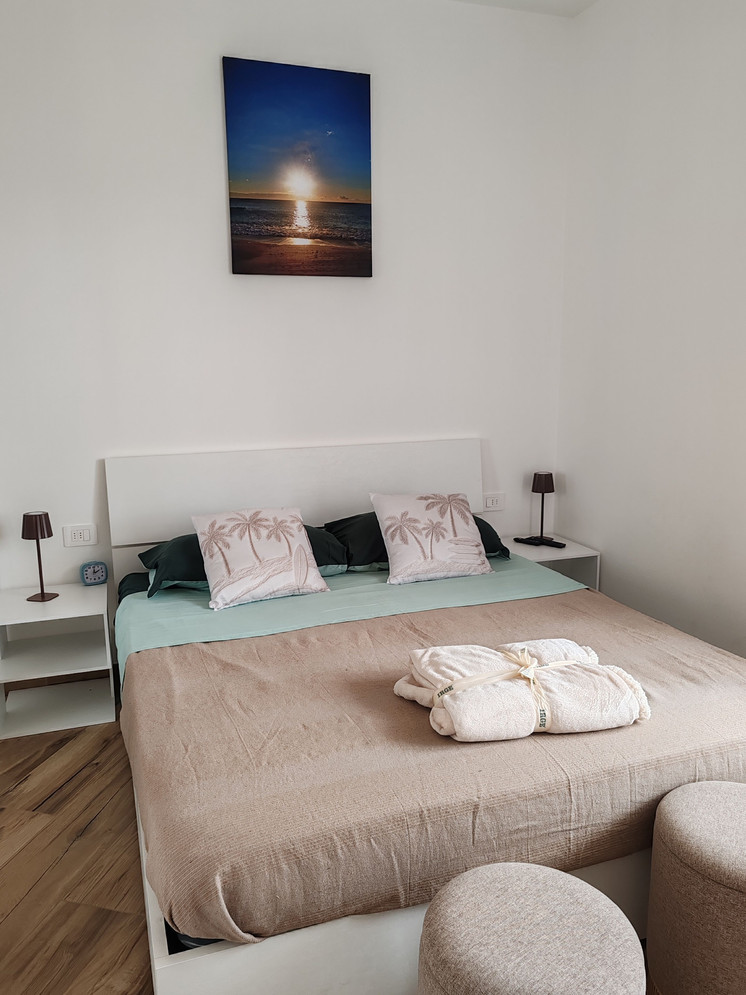 Una camera da letto ordinata con un letto, cuscini, un comodino con una lampada, un quadro appeso al muro e un pavimento in legno.