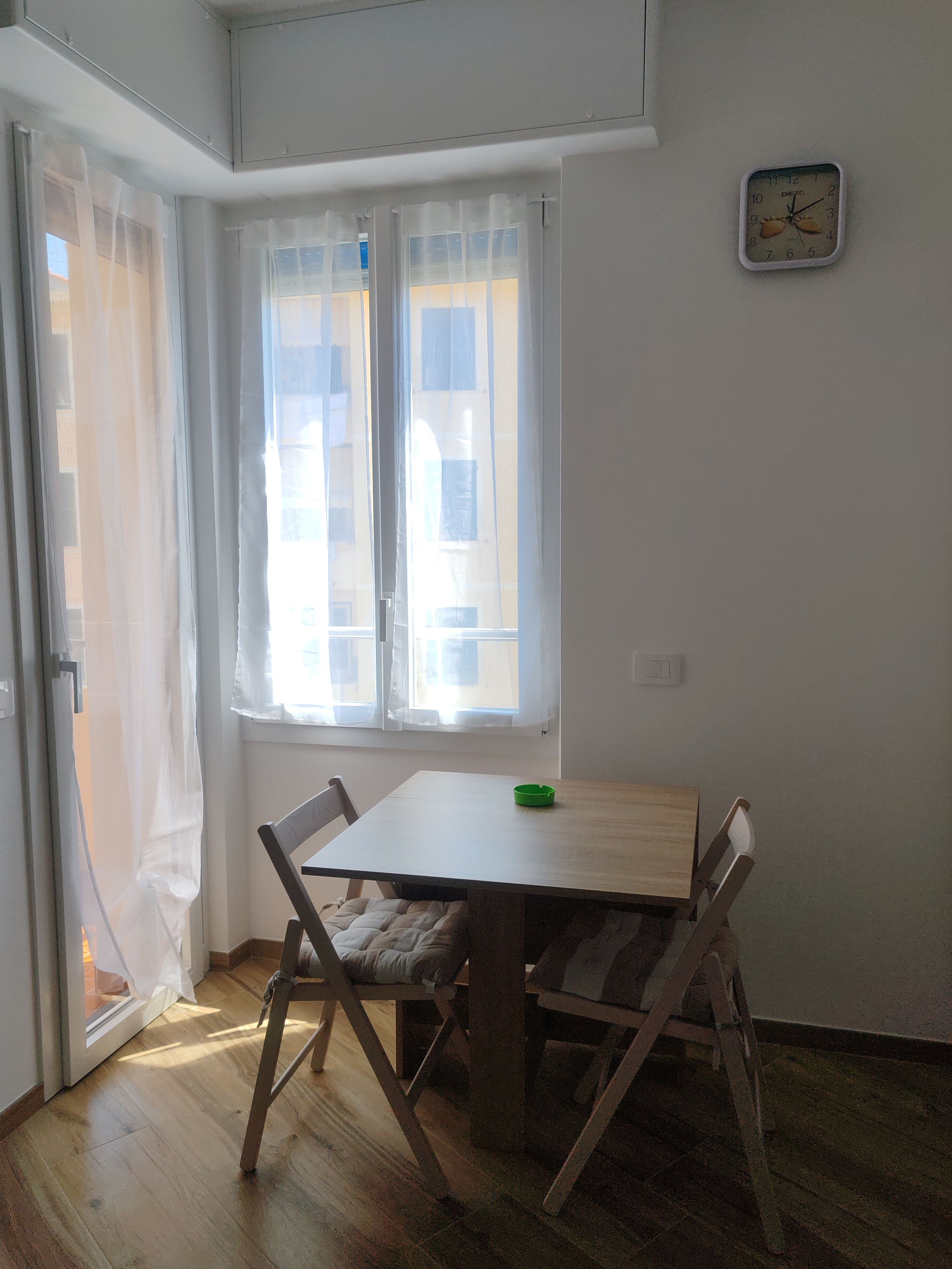 Ein Zimmer mit einem Holztisch, Stühlen, einem Fenster mit durchsichtigen Vorhängen und einer Wanduhr.