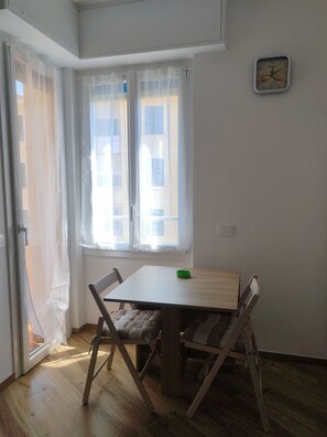 A room with a wooden table and chairs, a window with sheer curtains, and a wall clock.