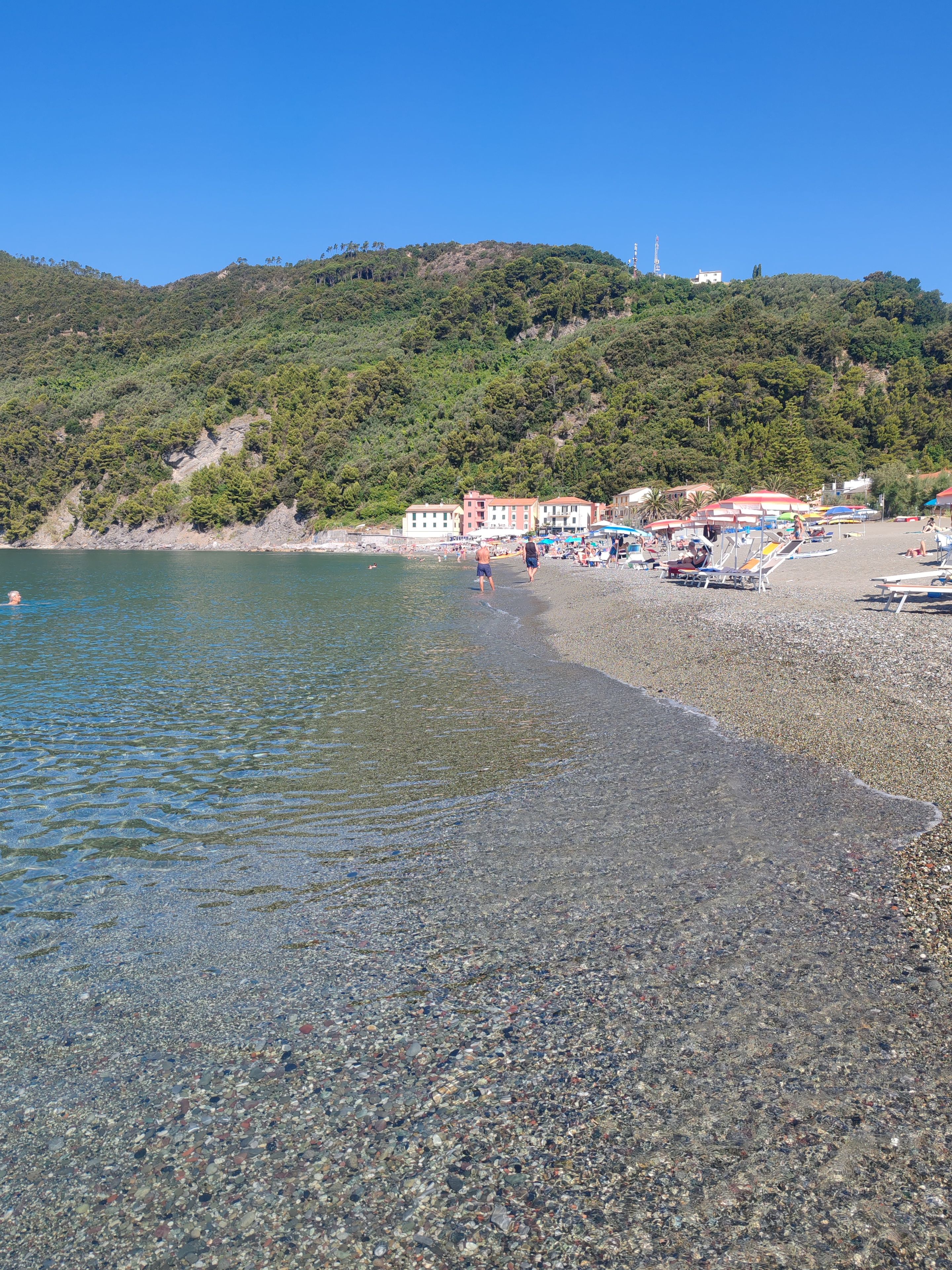 Ein Kiesstrand mit klarem Wasser, wenigen Personen und Gebäuden im Hintergrund.