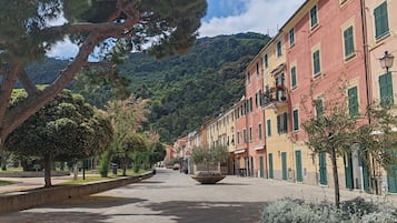 Une place pavée, bordée de bâtiments des deux côtés, avec un grand arbre au premier plan et une montagne en arrière-plan.