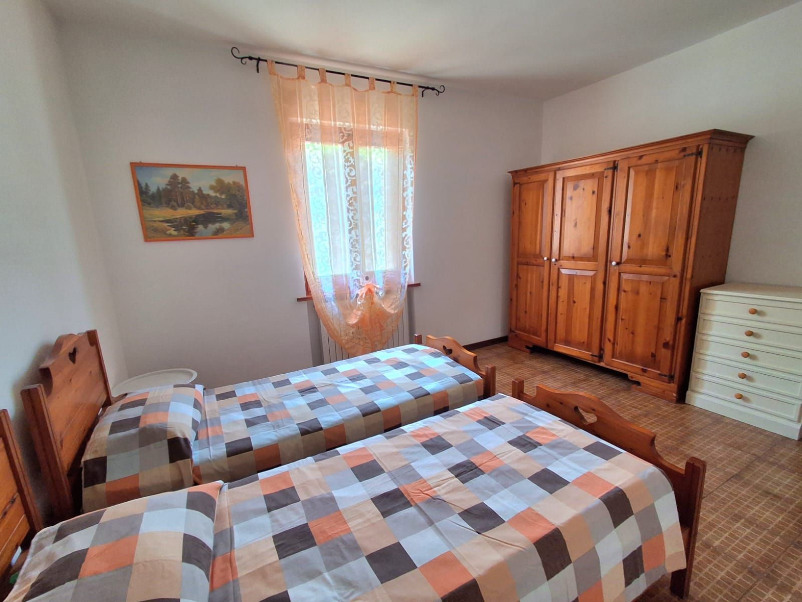 Quarto com duas camas de madeira, um guarda-roupa de madeira, uma cômoda branca pequena e uma janela com cortina translúcida.