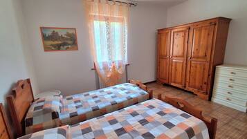 Quarto com duas camas de madeira, um guarda-roupa de madeira, uma cômoda branca pequena e uma janela com cortina translúcida.