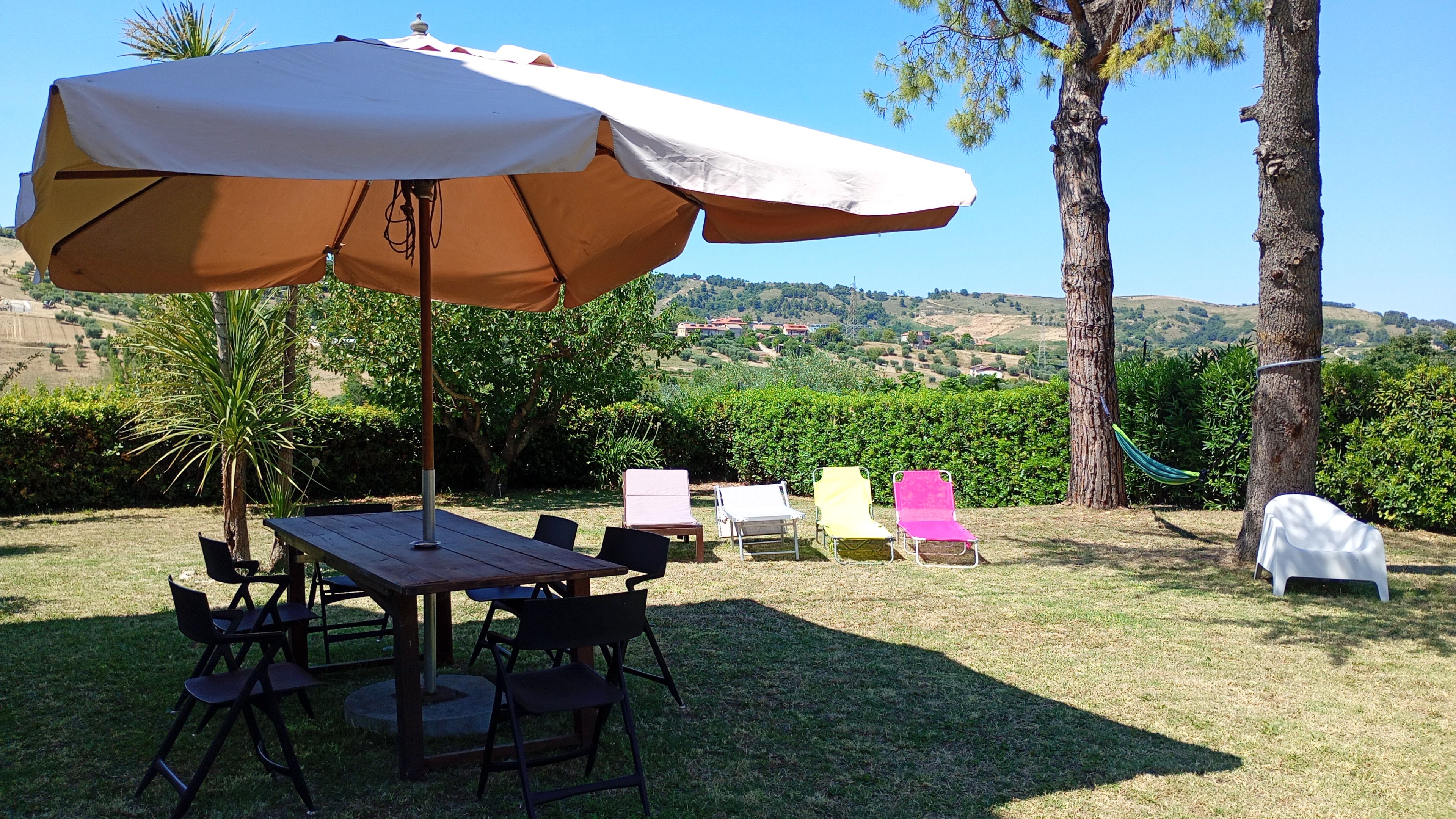 An outdoor patio with a table and chairs, a large umbrella, and a view of a hilly landscape.