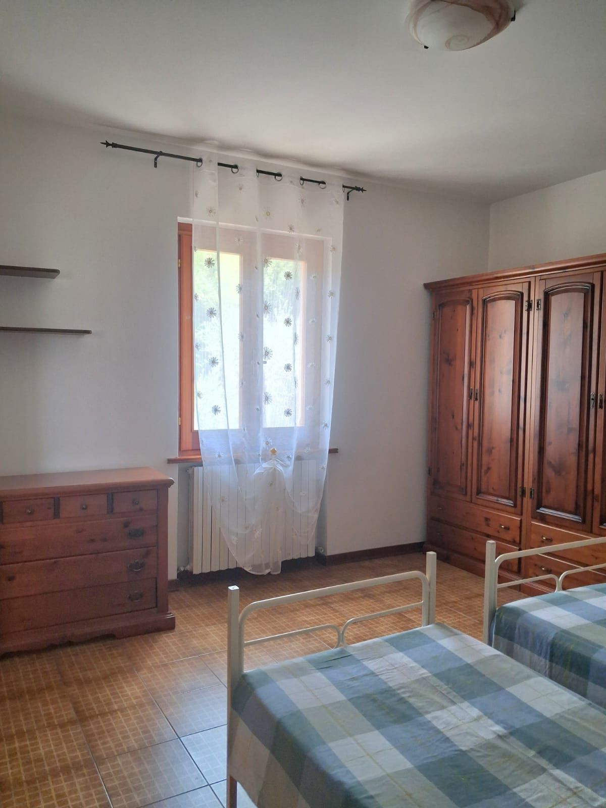 A bedroom with a wooden wardrobe, a bed with a checkered bedspread, a wooden dresser, and a window with a sheer curtain.