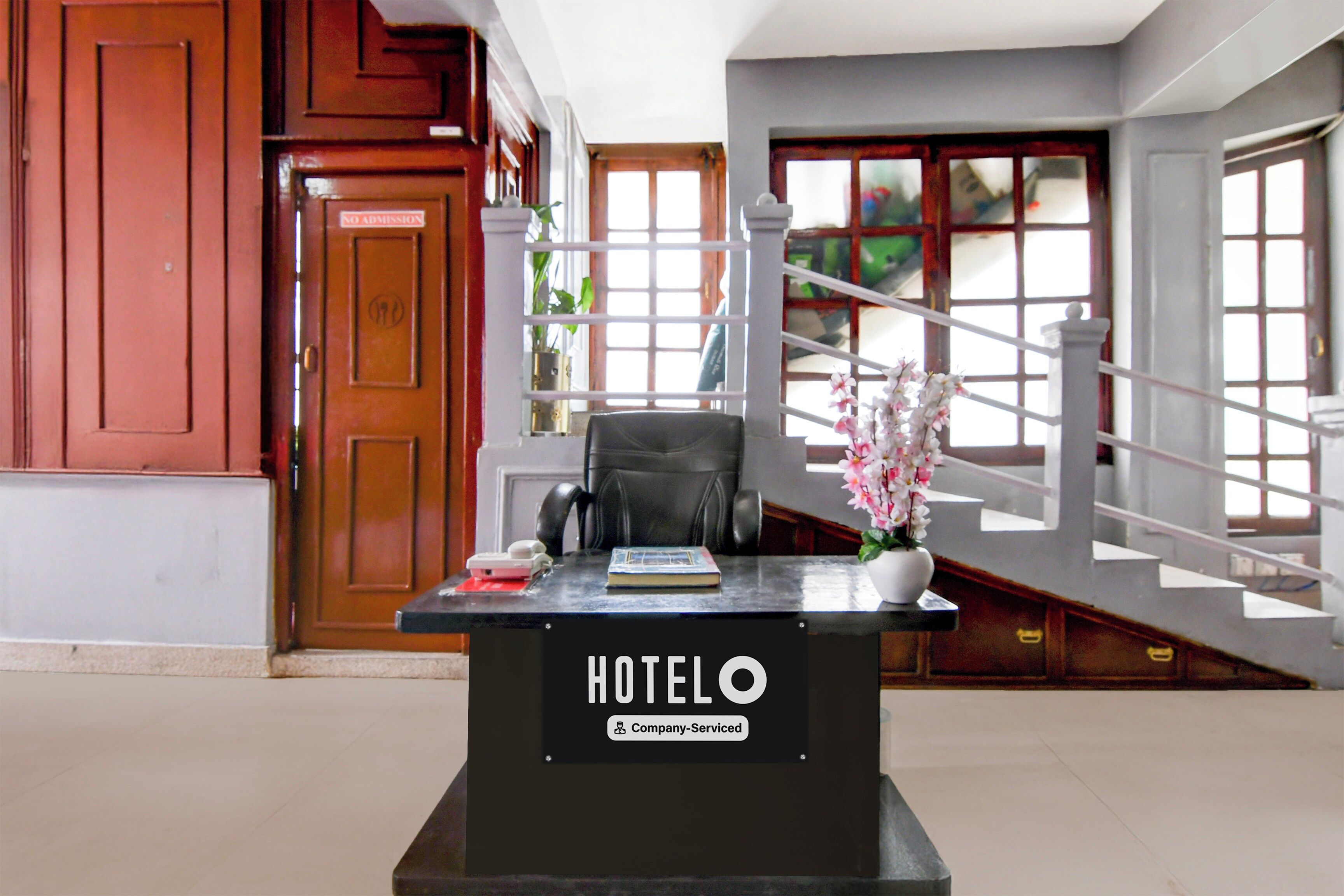 A hotel reception desk with a sign indicating it is company-serviced. There is a chair and a small table with a book and a vase of flowers.