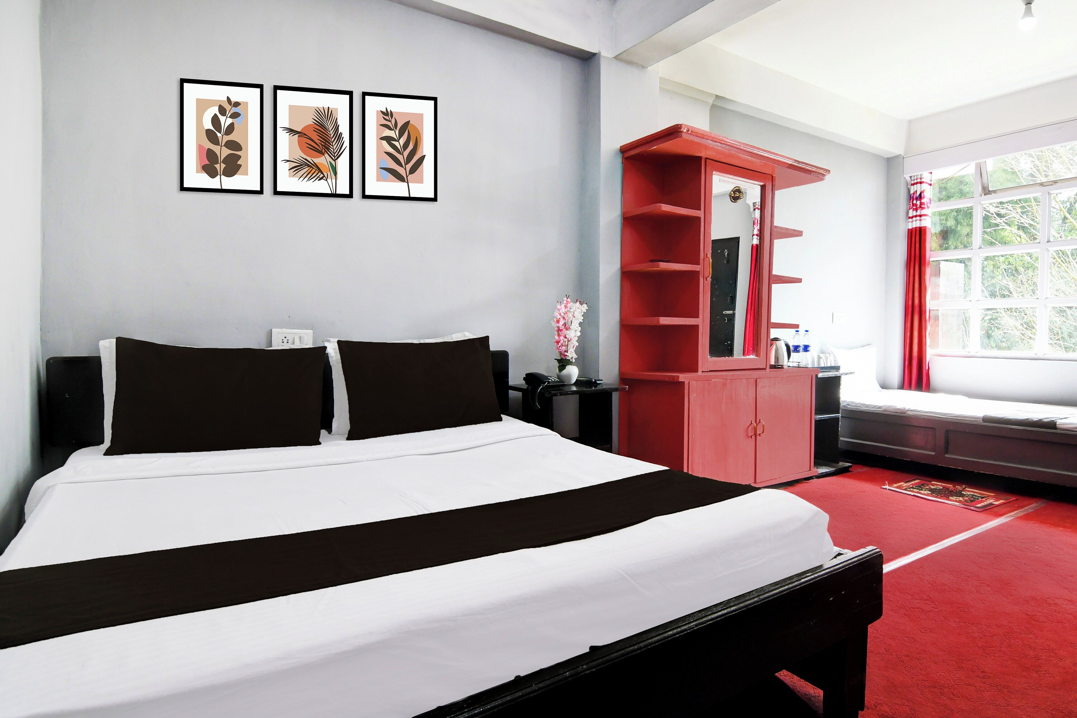 Une chambre d’hôtel avec un lit, une armoire rouge, une fenêtre donnant sur un beau paysage et trois tableaux encadrés accrochés au mur.