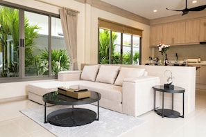A modern living room with a sofa, a coffee table, and a side table. There is a kitchen area with cabinets and a countertop visible in the background. - 2BR Villa Private Pool & Tropical Garden (Tambon Rawai)