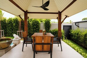 Een overdekt terras met een tafel en stoelen, omringd door groen en een decoratief vogelbeeldje.