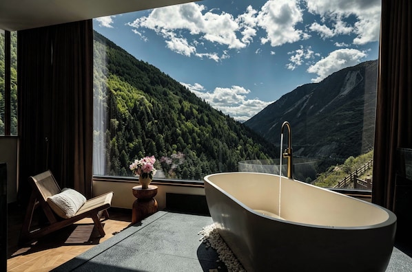 A room with a large window, a bathtub, a chair, and a vase with flowers.