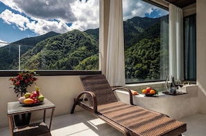 A balcony with a wicker lounge chair, a small table with fruit and a vase, and a view of a forested mountain range.