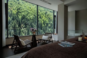 A bedroom with a large window offering a forest view, a bed with a magazine, a chair, and a small table with a vase of flowers.
