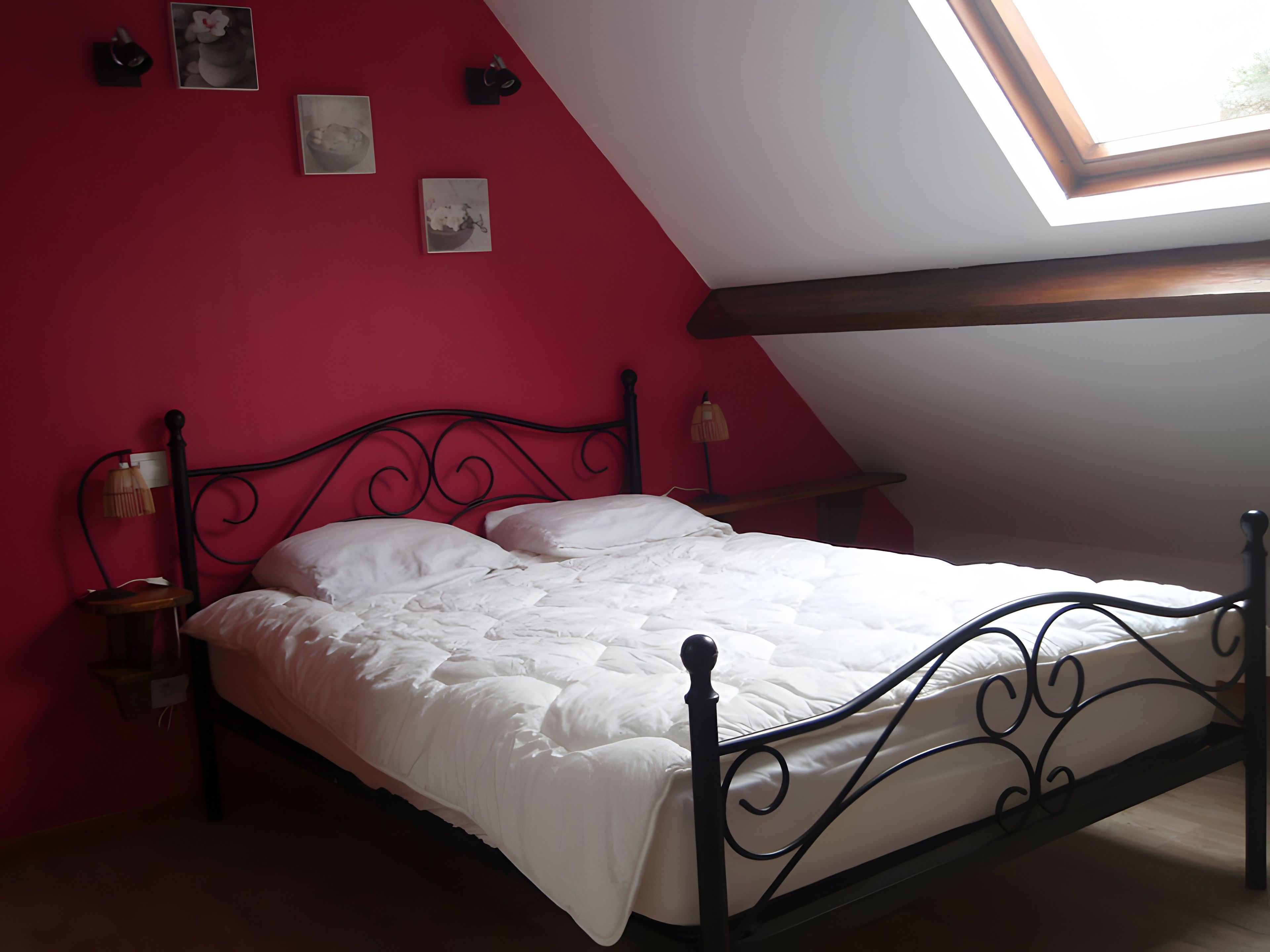 Un lit dans une chambre avec un plafond en pente et un puits de lumière, agrémenté d’un mur rouge et d’un cadre en métal noir.
