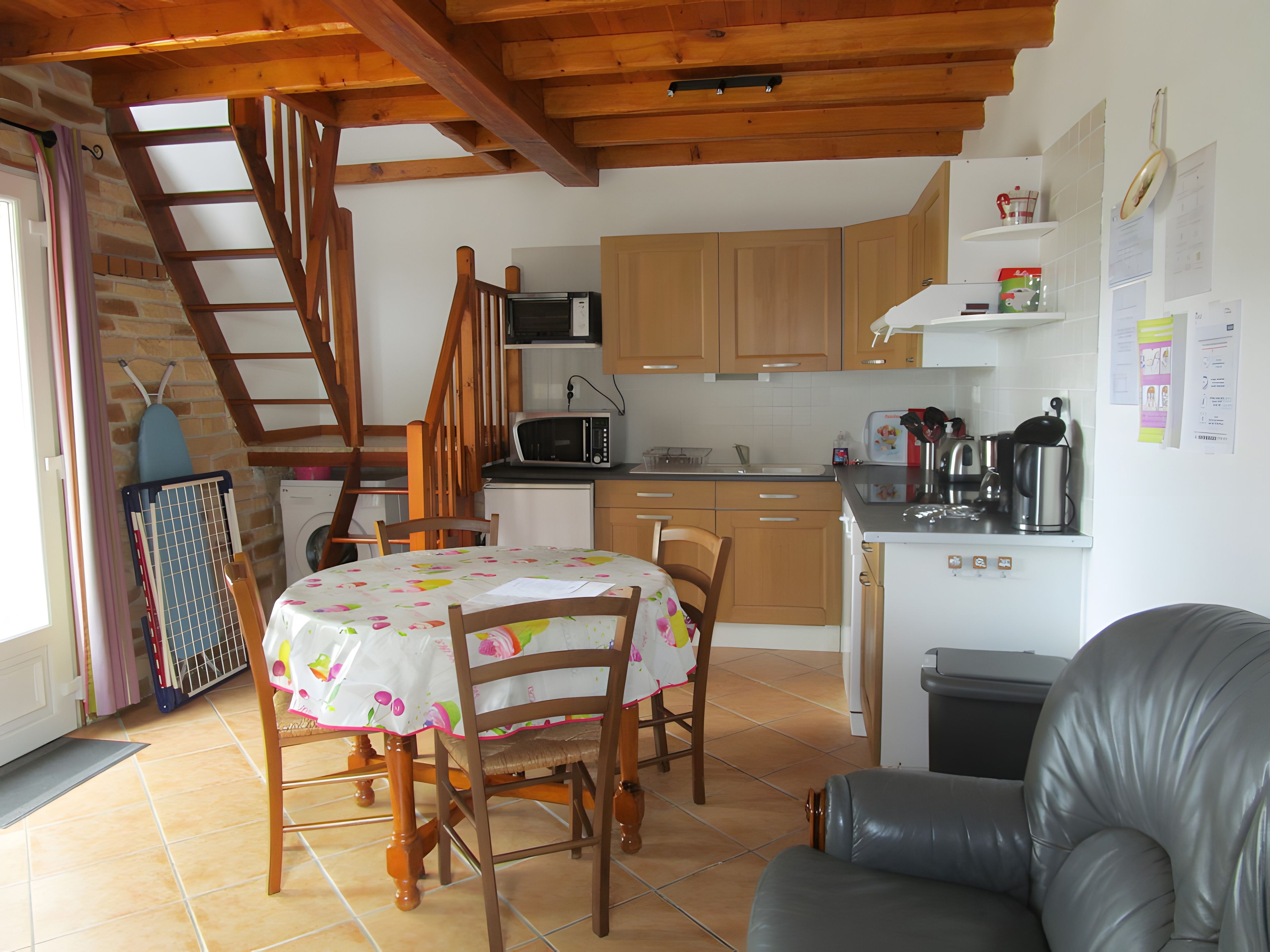 Une cuisine compacte avec des meubles en bois, un four à micro-ondes et un évier. On y trouve une table à manger avec des chaises et un canapé en cuir. Un escalier en bois mène à un niveau supérieur.