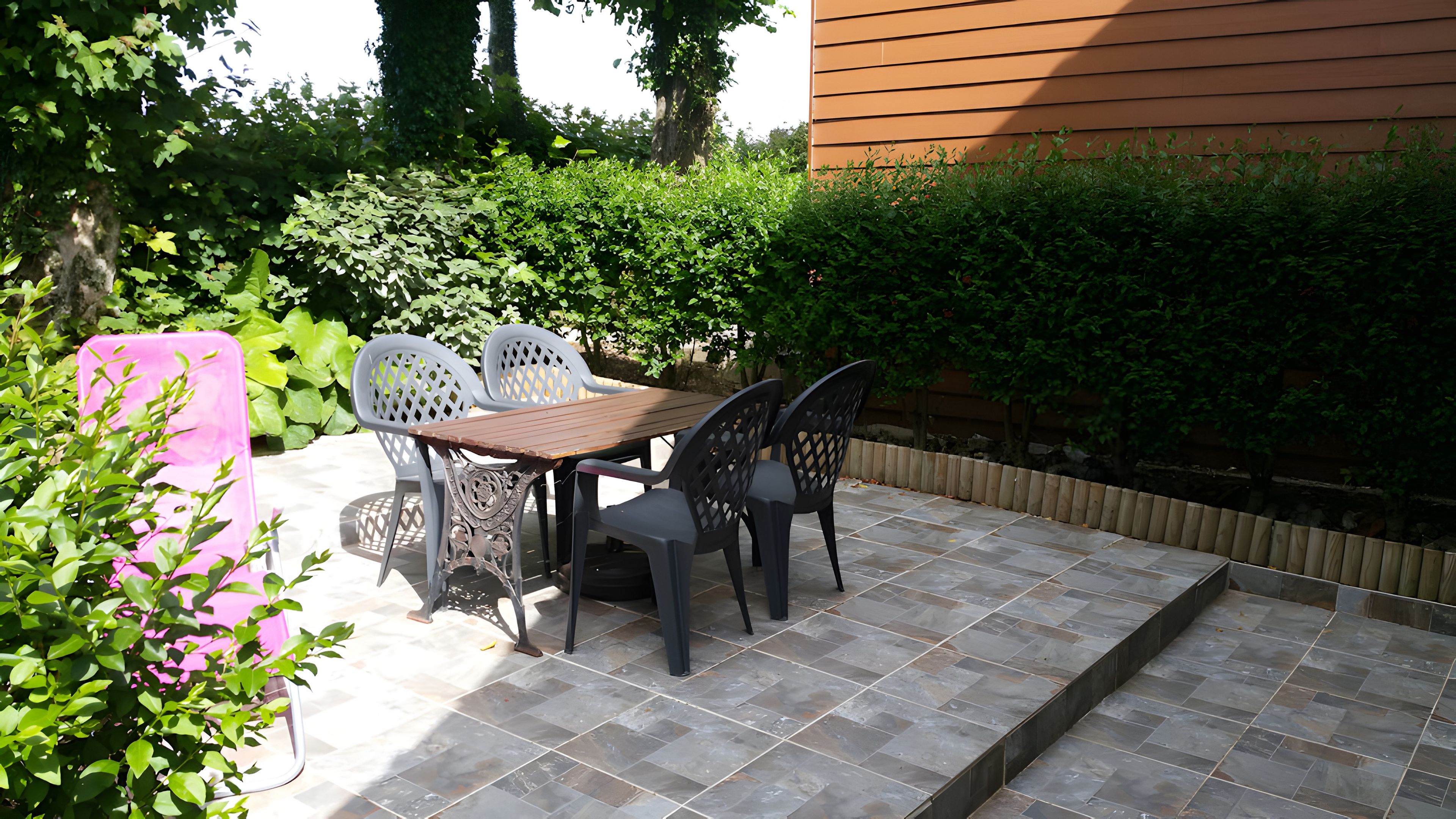 Une terrasse extérieure avec une table et des chaises en bois, entourée de verdure et d’une clôture en bois.