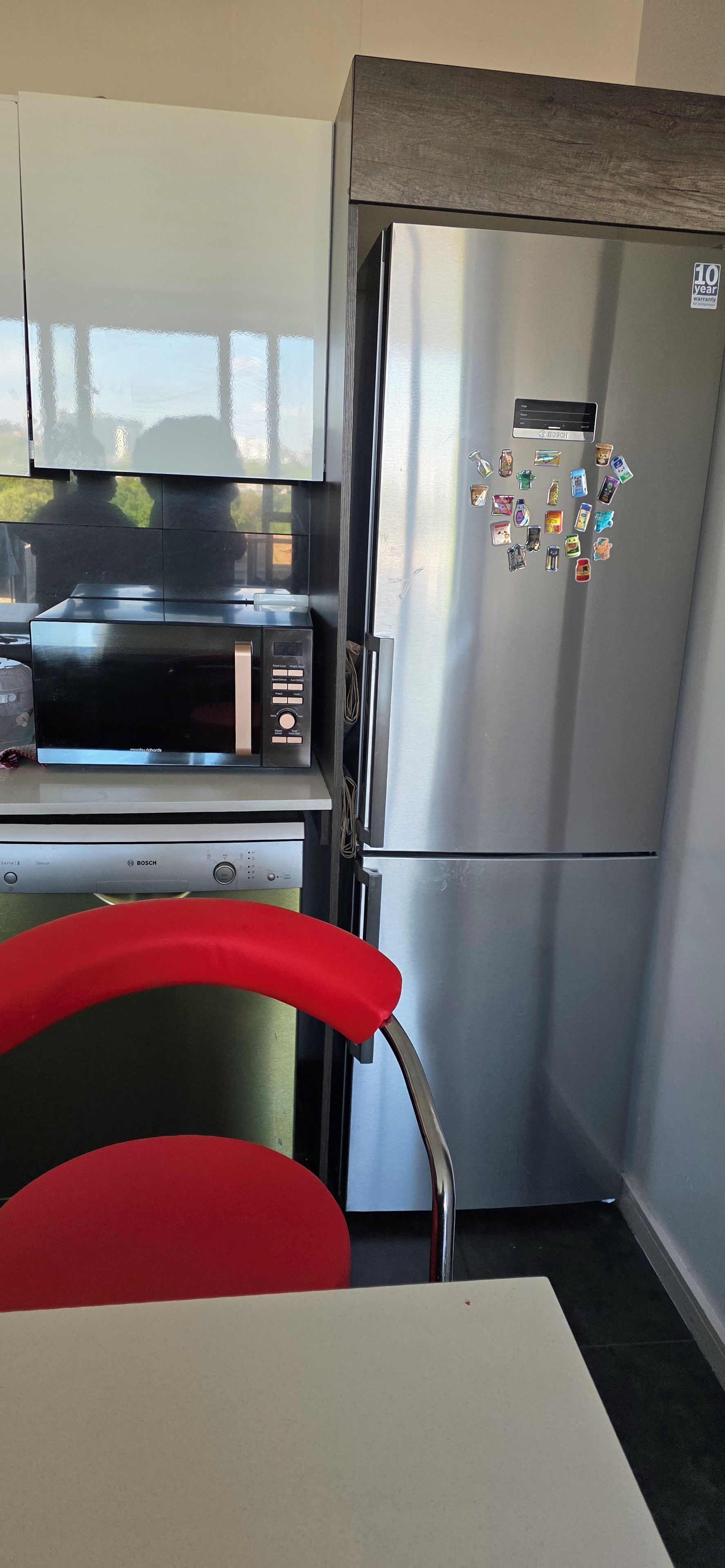 A compact kitchen with a stainless steel refrigerator, a microwave, and a red chair.