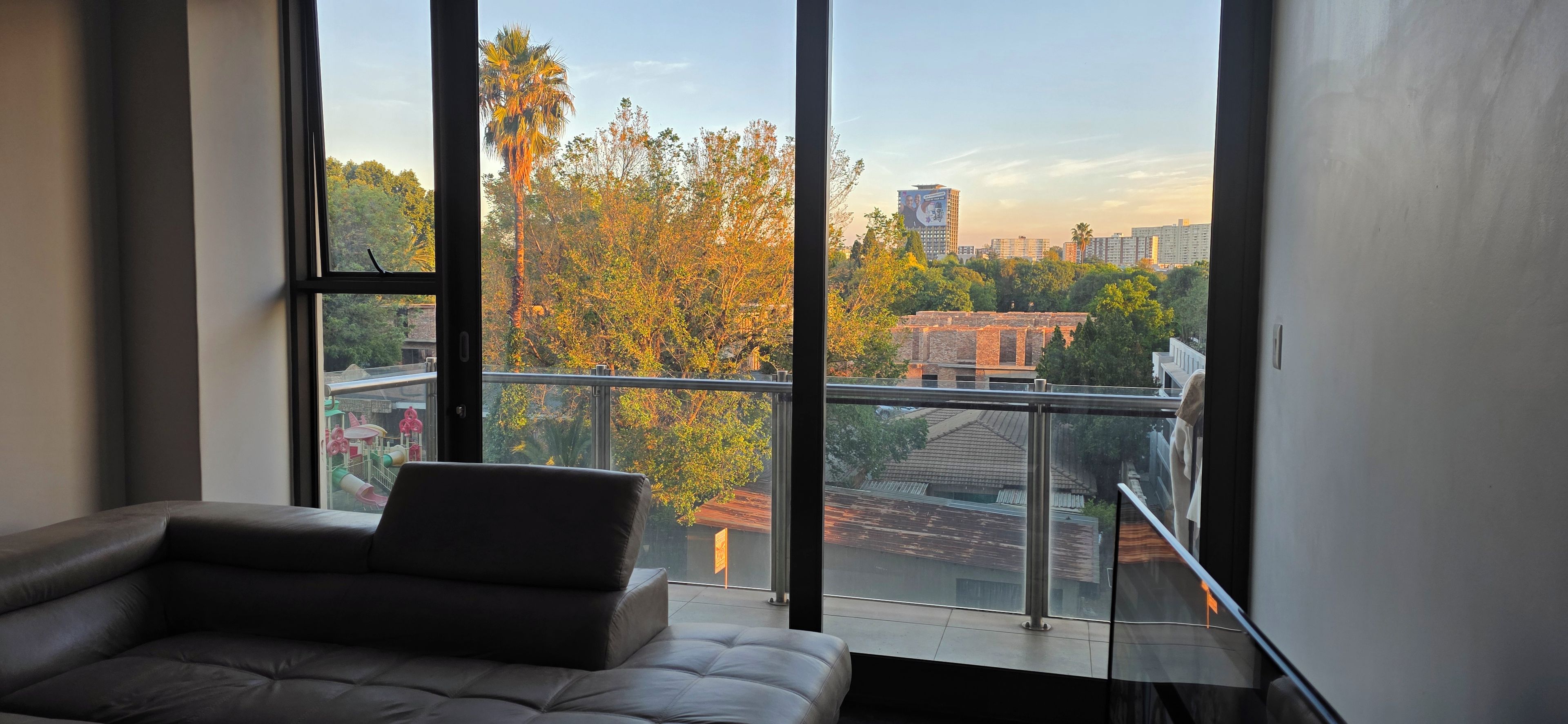Ein modernes Zimmer mit großem Fenster, einer Couch und einem Balkon mit Blick auf eine Stadtlandschaft mit Bäumen und Gebäuden.