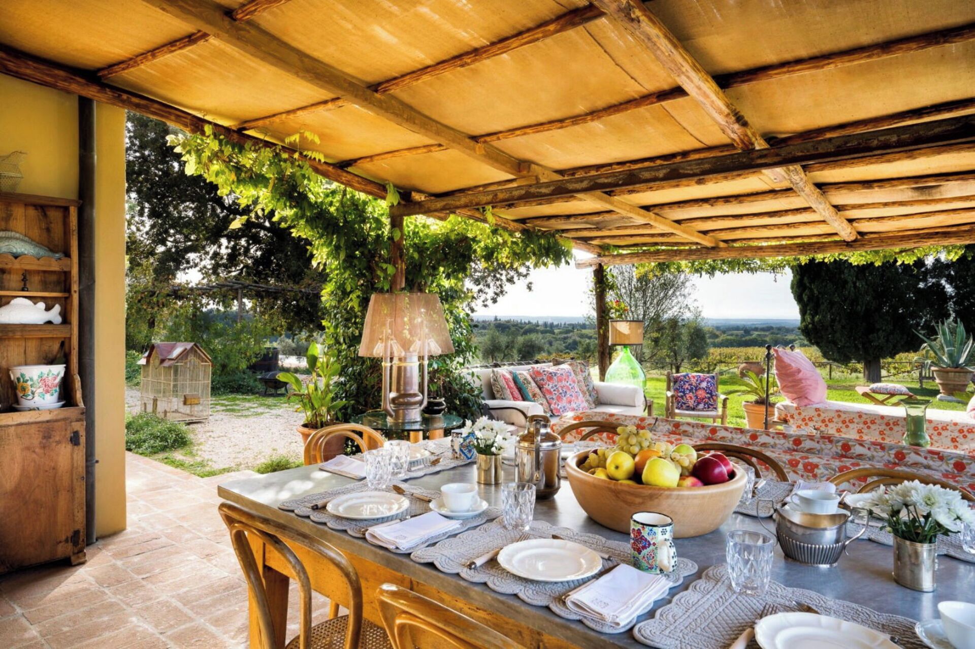 Un espace de restauration en plein air avec une table dressée pour quatre personnes, entourée de chaises, sous une pergola en bois offrant une vue sur la campagne.
