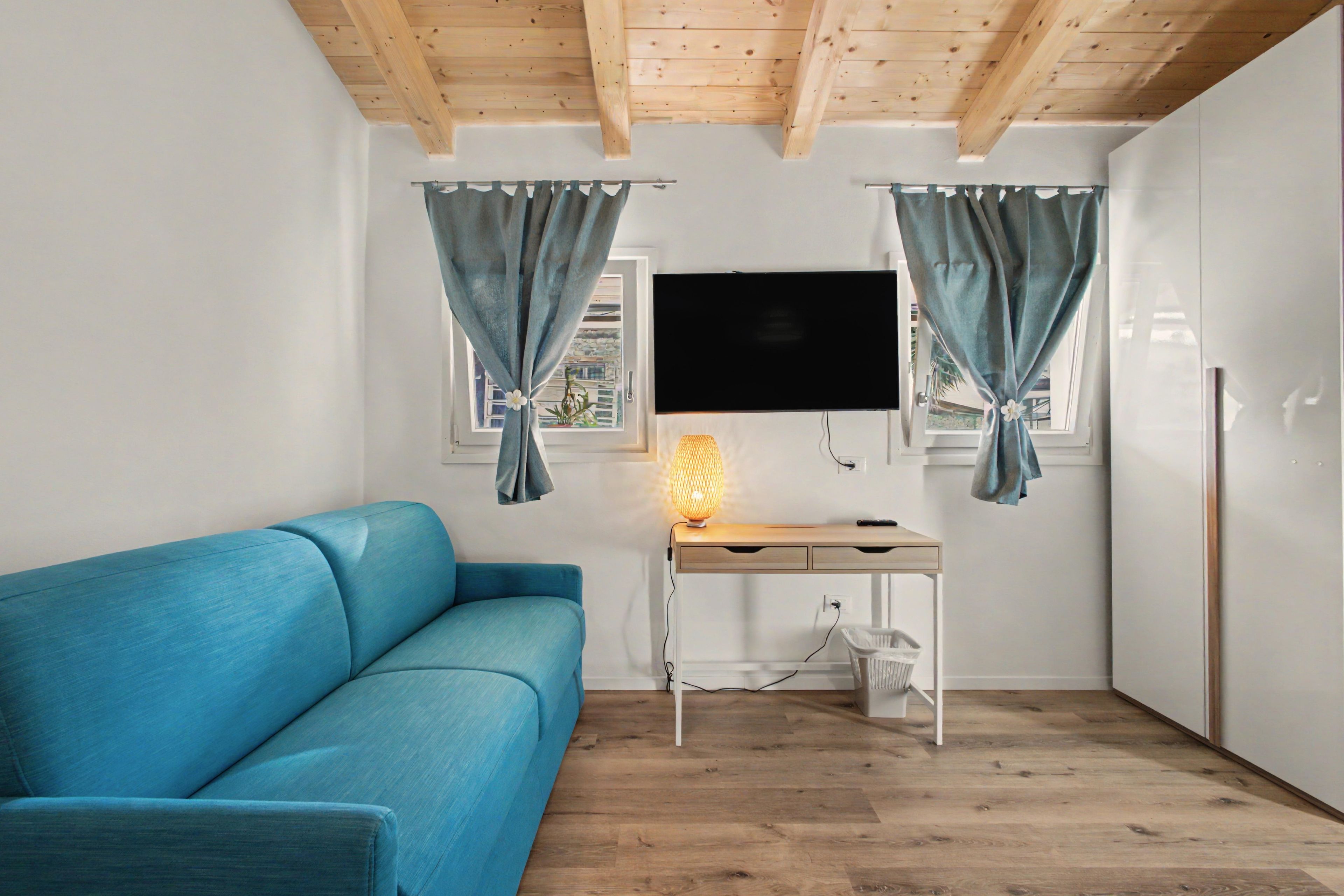 Una stanza con un divano blu, una scrivania in legno con una lampada, una TV a parete e una finestra con tende.