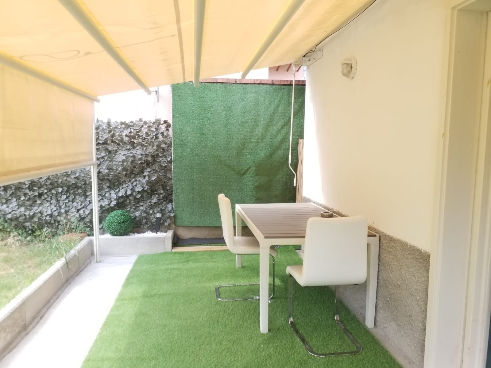 Un piccolo balcone con un tavolo e due sedie, un pavimento di erba sintetica verde e pareti, una bianca e l'altra in pietra, sullo sfondo.