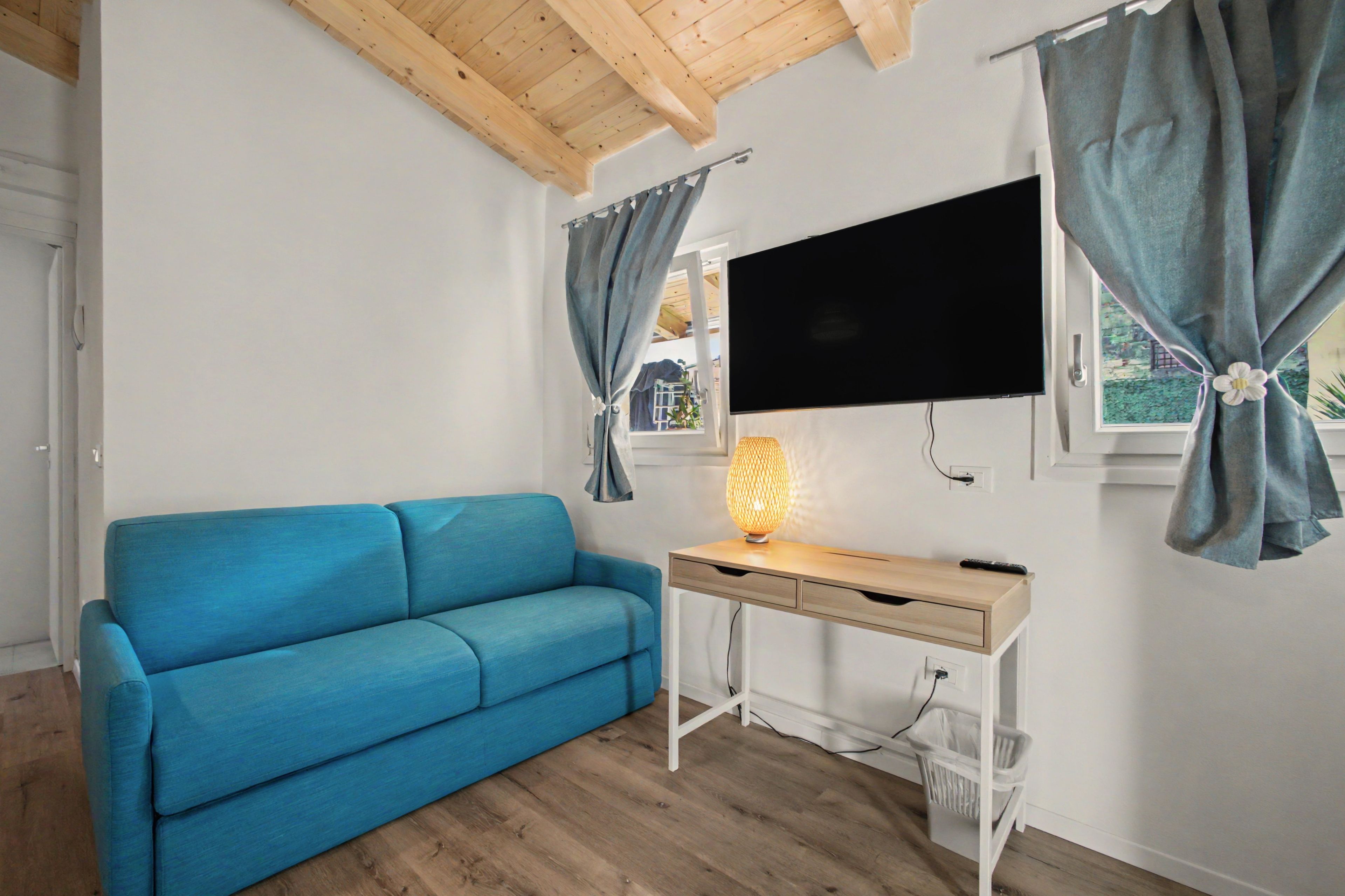 Quarto com sofá azul, escrivaninha de madeira, TV fixada na parede e janela com cortinas.