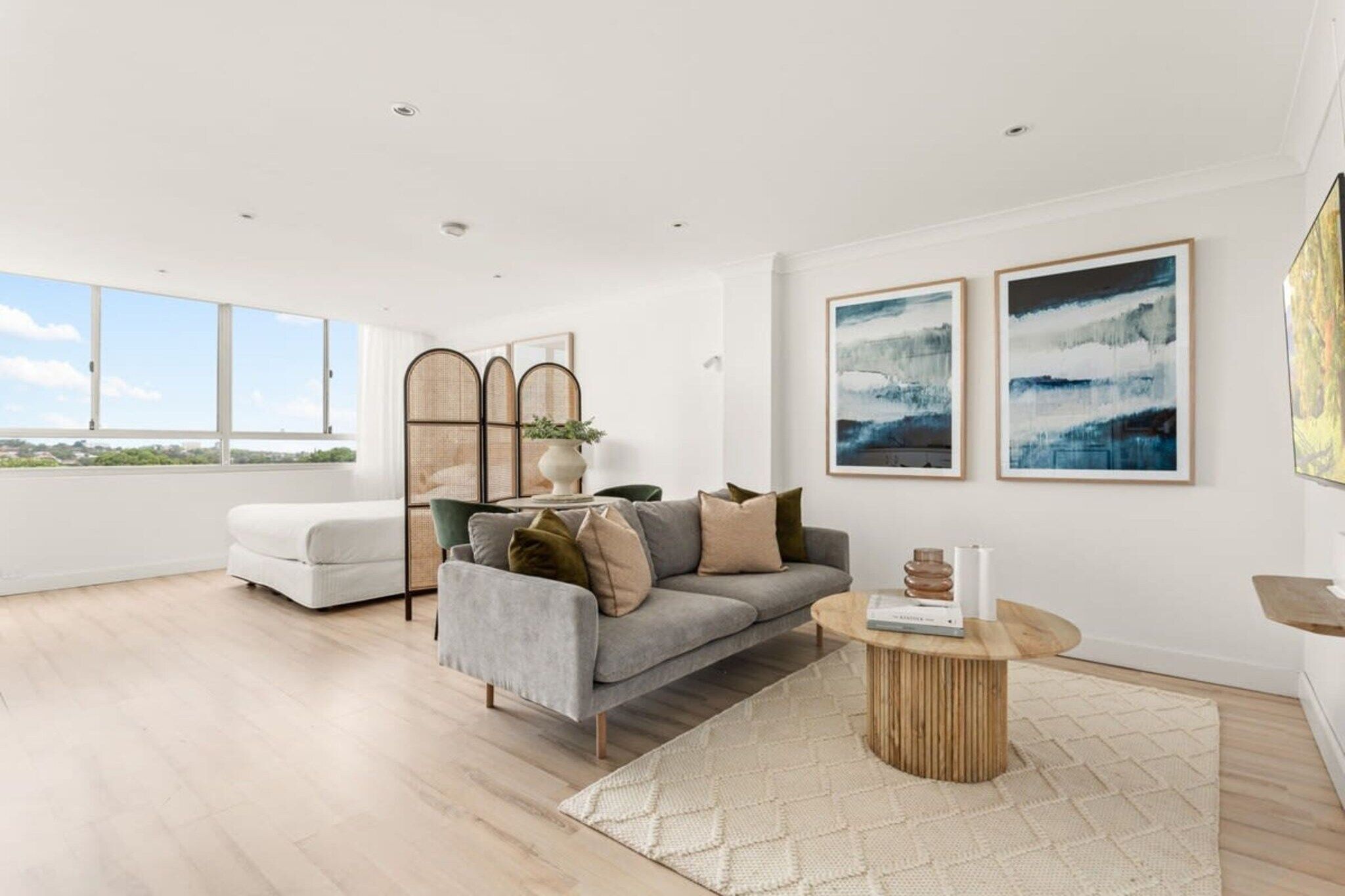 A modern living room with a grey sofa, a white armchair, and a round wooden coffee table. There are two framed abstract paintings on the wall and a large window providing a view of the outdoors.
