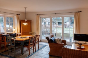 Ein Essbereich mit einem Holztisch und Stühlen, einem Fernseher auf einem Schrank und Blick ins Freie durch große Fenster.