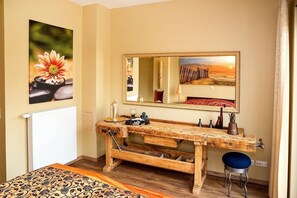 A room with a wooden vanity, a large mirror, a bed with patterned bedding, and a painting of a flower. - Vrbo Property (Lenz)