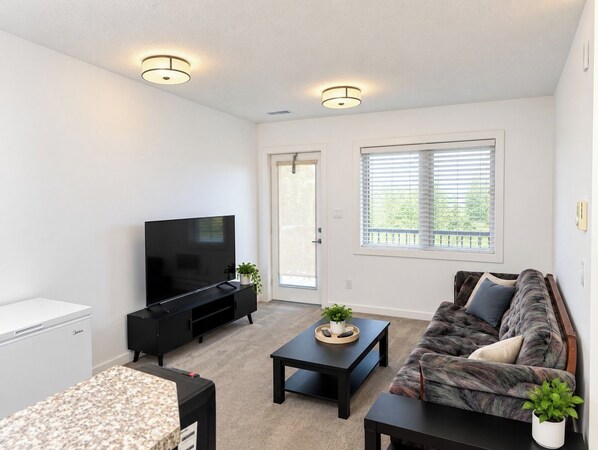 A modern living room with a flat-screen TV, a sofa, a coffee table, and a small kitchen area with a refrigerator.