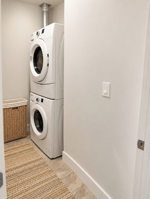A laundry room with stacked front-loading washers and dryers, a wicker basket, and a beige rug. - Spacious Yorkton Apartment (Yorkton)