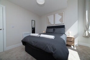 A modern bedroom with a large bed, a bedside table with a lamp, a wall-mounted mirror, and two framed artworks. - Sovereign Quay 1BR Apartment | Old Town Hull (Kingston upon Hull)