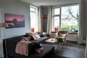 A living room with a sofa, armchair, and coffee table, large windows with curtains, and a painting on the wall. - Nature house "De Blauwe Reiger (Slootdorp)