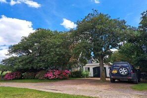 A residential driveway with a parked SUV, a house with a dark-colored garage, and lush greenery including flowering bushes. - Nature house "De Blauwe Reiger (Slootdorp)