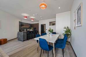 Een moderne eethoek met een witte tafel en blauwe stoelen, een keuken op de achtergrond en een potplant aan de rechterkant.
