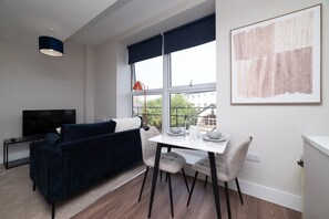 Ein modernes Wohnzimmer mit einem dunkelblauen Sofa, einem Esstisch mit Stühlen, einem Fernseher und einem großen Fenster mit Blick auf Gebäude und Grünflächen.
