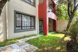 A modern two-story building with a red and beige facade, a stone pathway leading to the entrance, and a small garden area with grass and shrubs. - Elegant 2-Bedroom Retreat in Fourways, Johannesburg (Sandton)