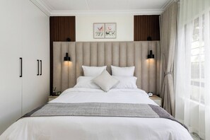 Un letto rifatto con lenzuola bianche e una coperta grigia, affiancato da due comodini con lampade, una sveglia e un piccolo oggetto decorativo.
