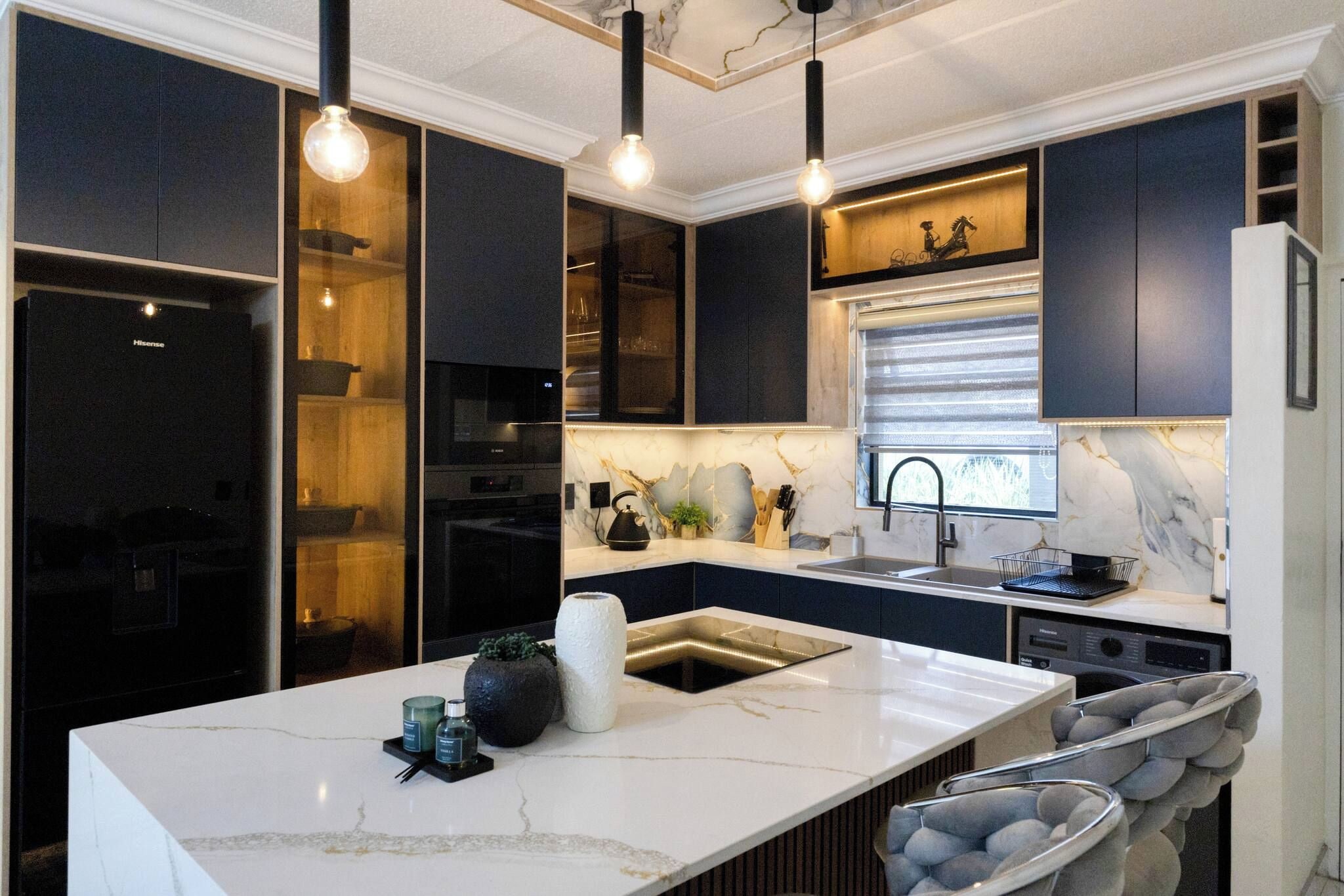 Une cuisine moderne avec des meubles de couleur sombre, un îlot en marbre, des luminaires suspendus et un four encastré.