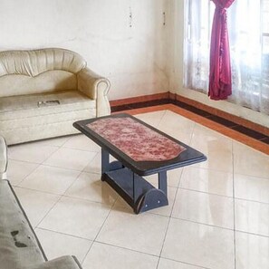 Une pièce avec un canapé beige, une petite table sur laquelle repose un coussin rouge, et une fenêtre avec un rideau.