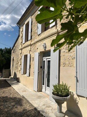 Un bâtiment en pierre de deux étages, doté de volets blancs, d’une plante en pot et d’un chemin pavé.