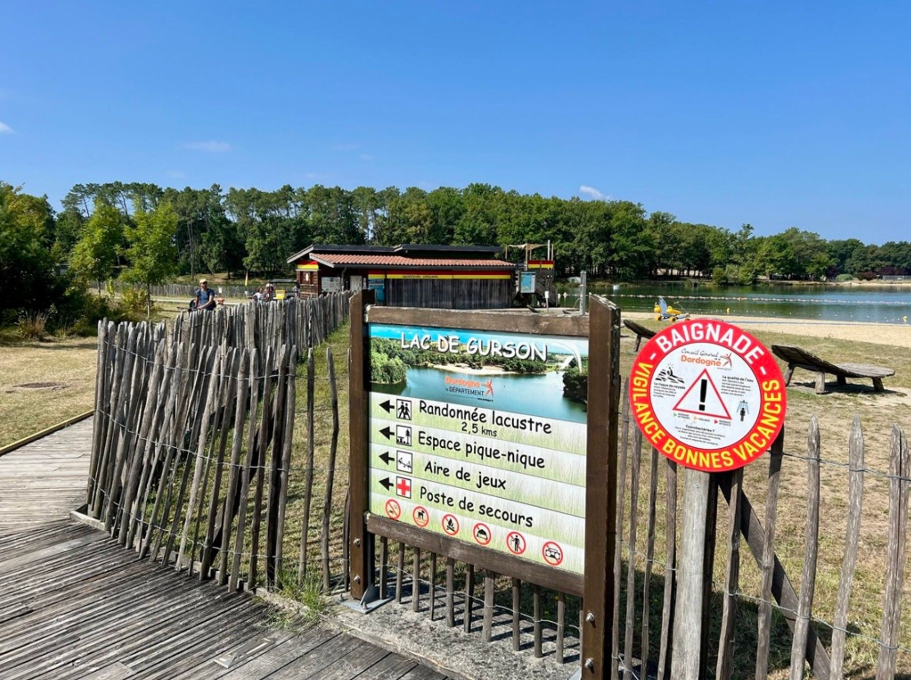 A wooden boardwalk leading to a beach area with a sign indicating facilities such as a picnic area, game area, and a post for resting.
