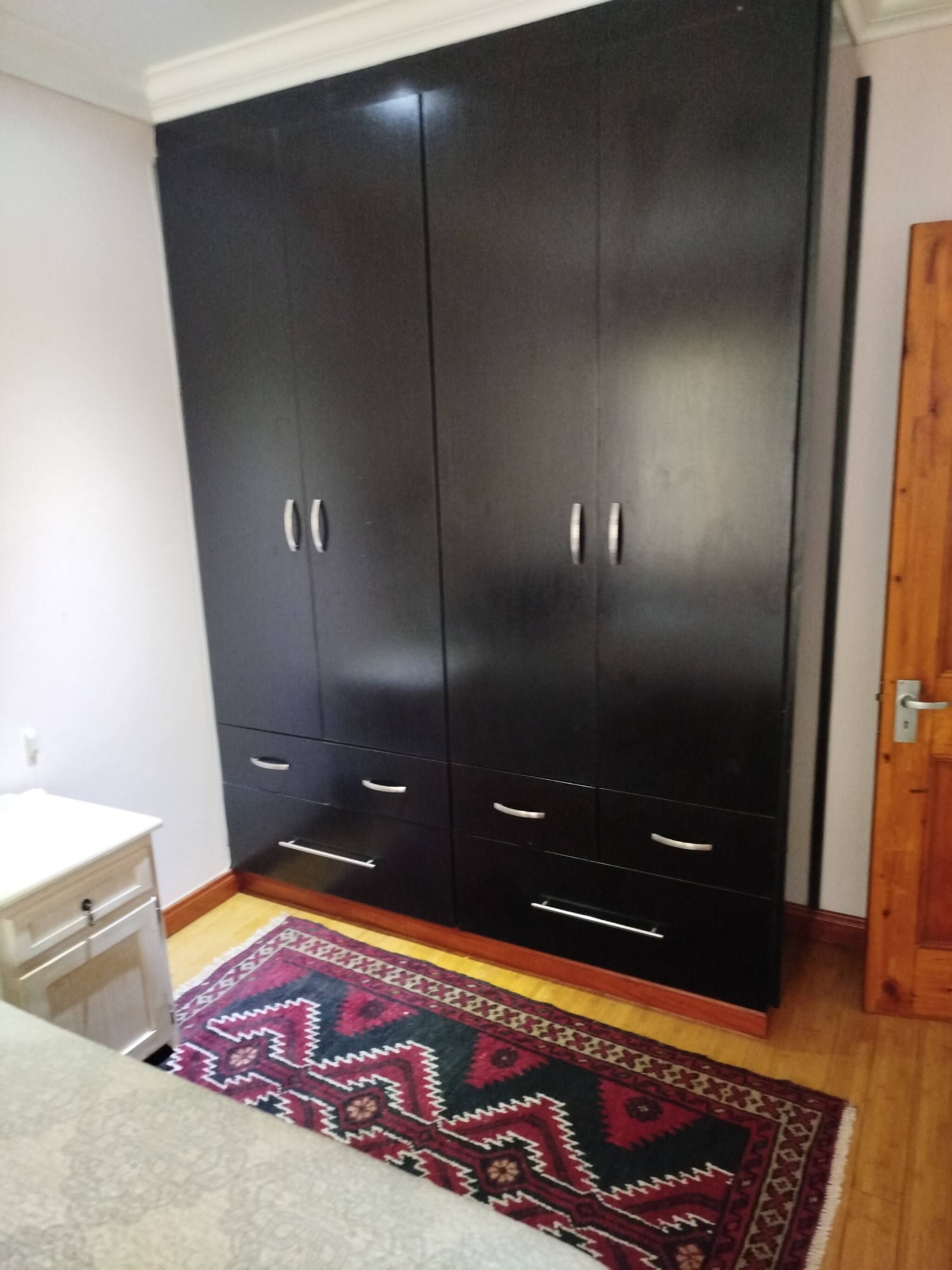 Una habitación con un armario negro grande, una mesita de noche blanca y una alfombra estampada en el suelo.