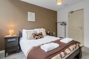 Een net opgemaakt bed met witte lakens en een bruine deken, een nachtkastje met een lamp, een lijst met een foto aan de muur, en een kapstok.