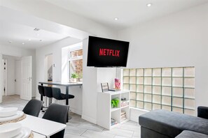 Een moderne woonkamer met een wandtelevisie waarop 'NETFLIX' staat, een eethoek met een tafel gedekt voor vier personen, en een zithoek met een bank.