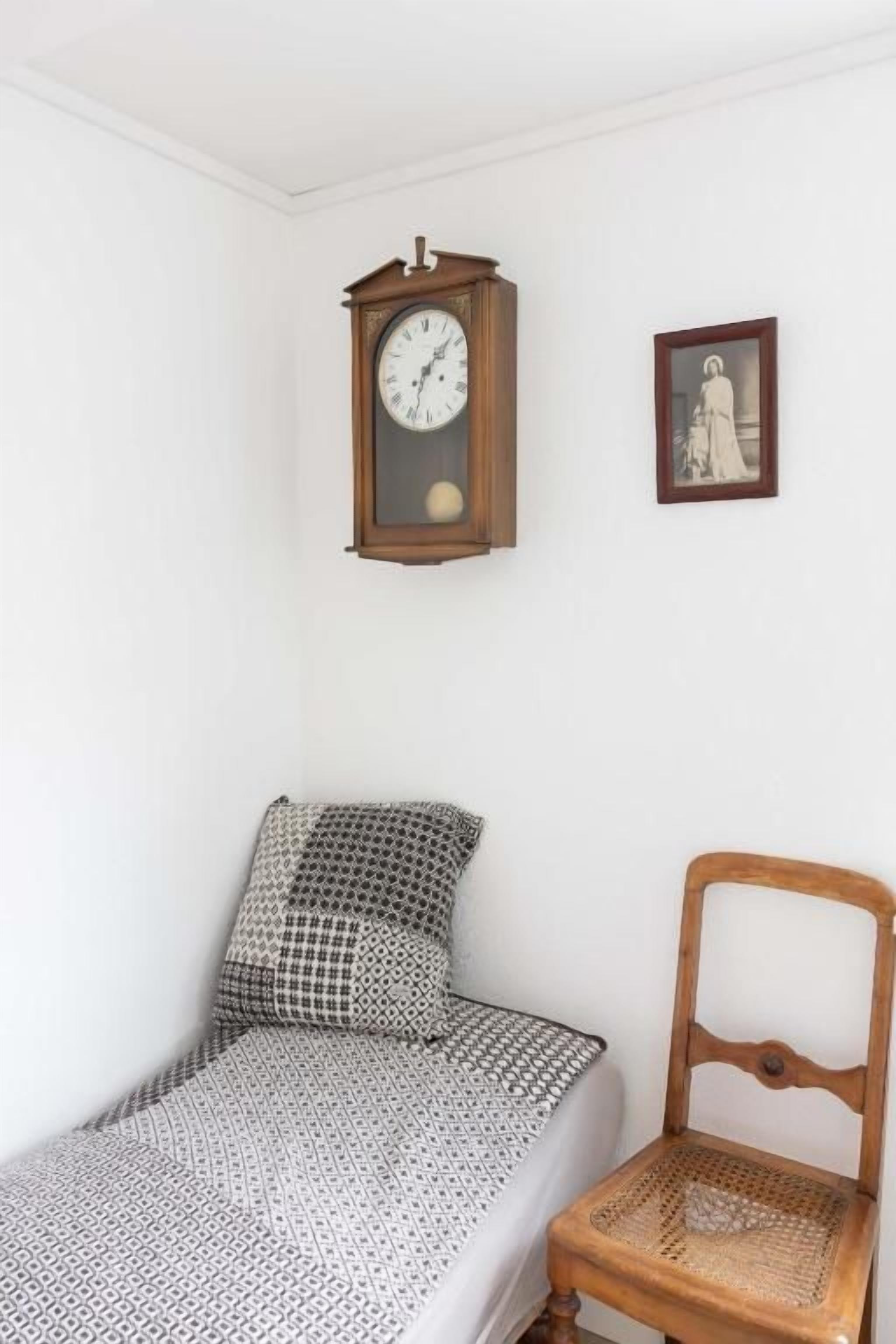 A small room with a single bed, a wooden chair, a wall clock, and a framed picture.