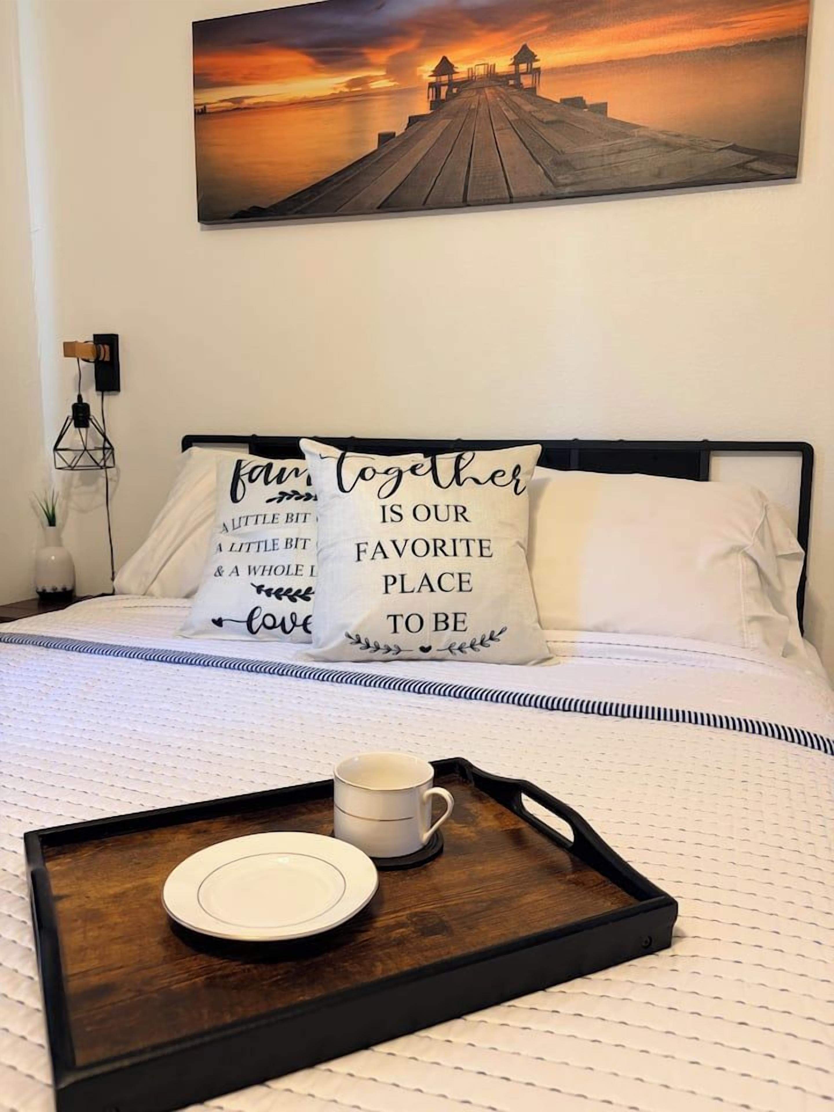 A neatly made bed with a tray of a cup and a plate, a painting of a sunset on the wall, and two pillows with a family-themed message.