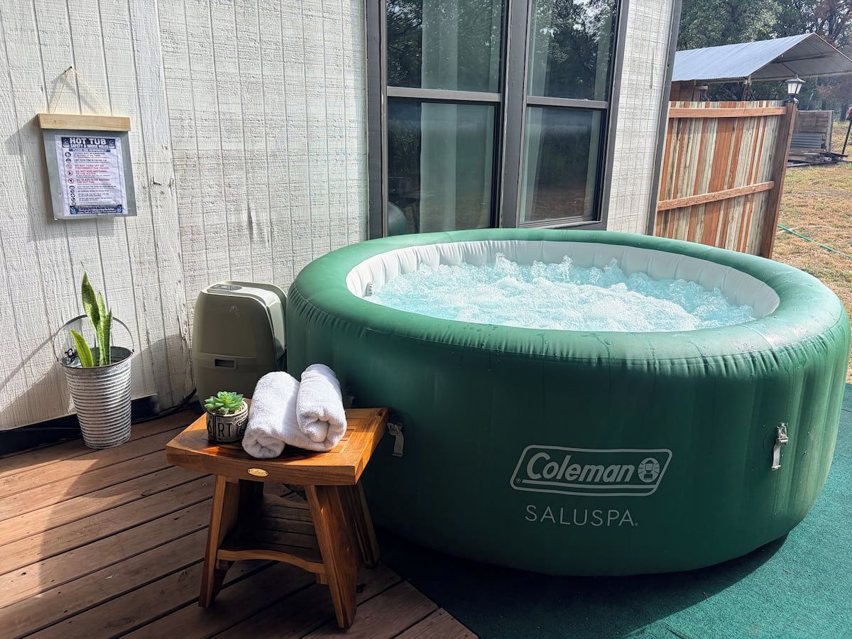 A green inflatable hot tub with a Coleman logo, a wooden stool with towels, and a metal bucket with plants.