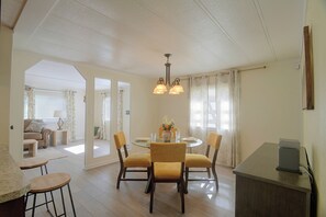 Ein Essbereich mit einem runden Glastisch, vier Stühlen und einer Chandelierleuchtre. Im Hintergrund befindet sich eine Couch und ein kleiner Tisch.