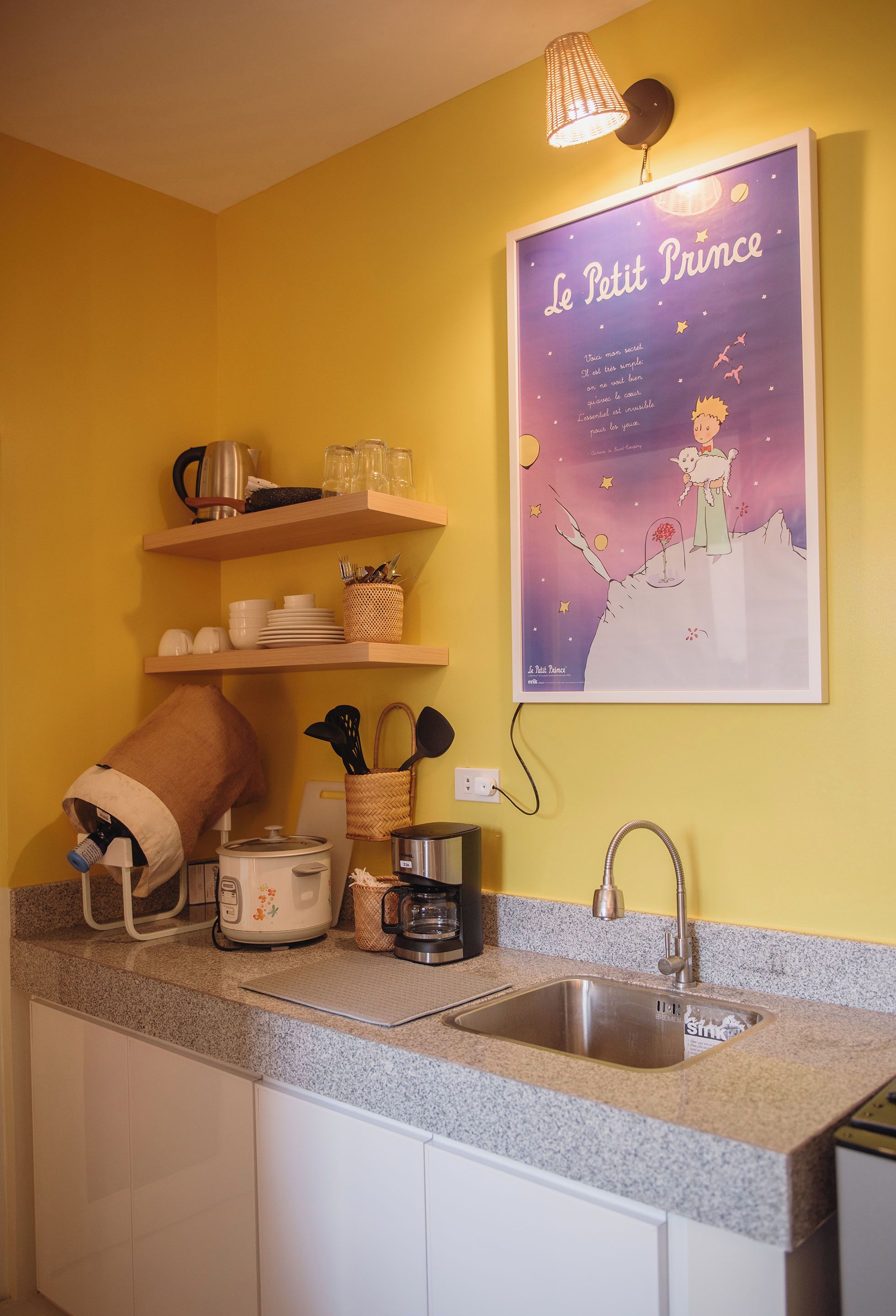 Une cuisine dotée d’un plan de travail en granit, d’un évier en acier inoxydable, d’une bouilloire, d’un grille-pain et d’une affiche du « Petit Prince » accrochée au mur.