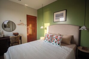 Un dormitorio con una cama, una puerta de madera, un espejo, un cuadro enmarcado en la pared y una mesita con una silla.